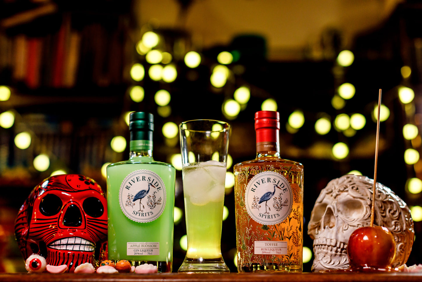 Two bottles of Riverside Spirits (Apple Blossom Gin Liqueur and Toffee Rum Liqueur) with a cocktail on a bar counter, surrounded by decorative skulls and fruits for Halloween.