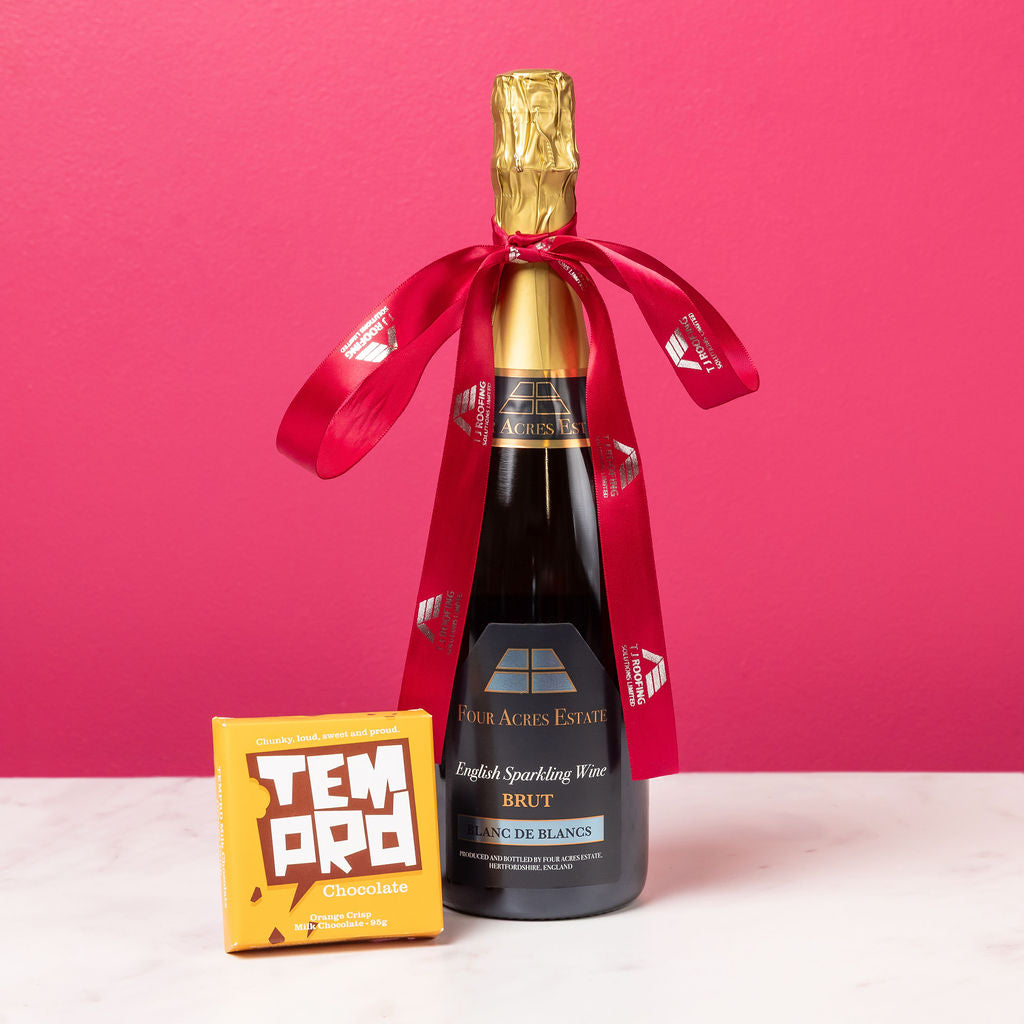 Bottle of Bottle of Four Acres Estate English Sparkling Wine Rose with a red ribbon and Temprd chocolate bar against a pink background