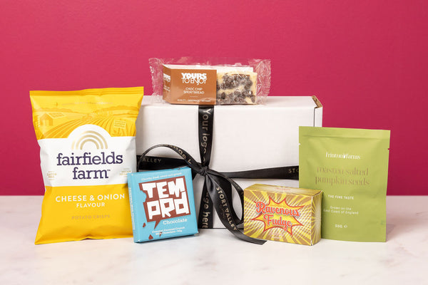 Personalised Stort Valley Gift box with assorted food products including a cheese and onion packet, a tea box, and other snacks on a pink background.