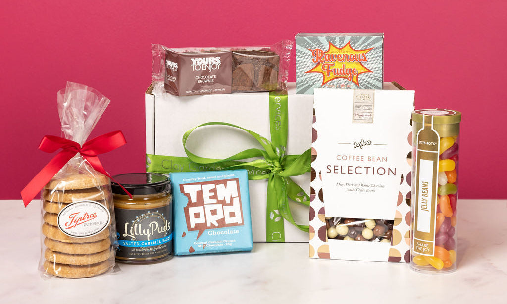Assorted gift items including cookies, tea, and coffee beans on a pink background