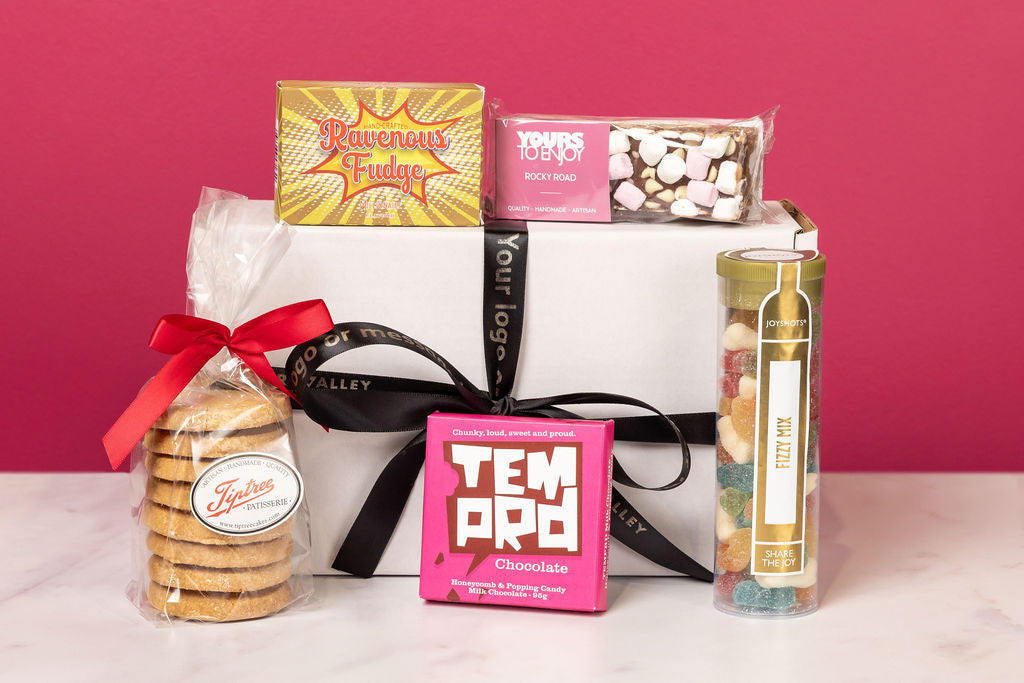 Personalised Stort Valley gift box with assorted snacks and treats including cookies, chocolate bars, and a tube of candy on a pink background.