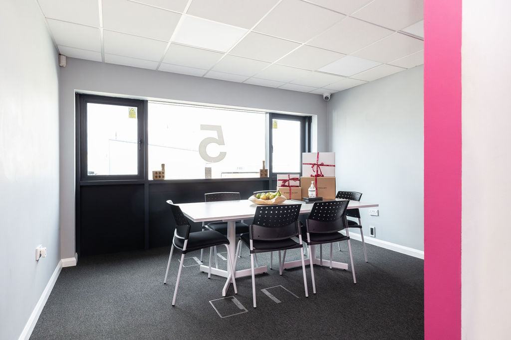 Modern office space with a table and chairs, Stort Valley Gifting branded personalised hampers, large windows, and a pink accent on the right.