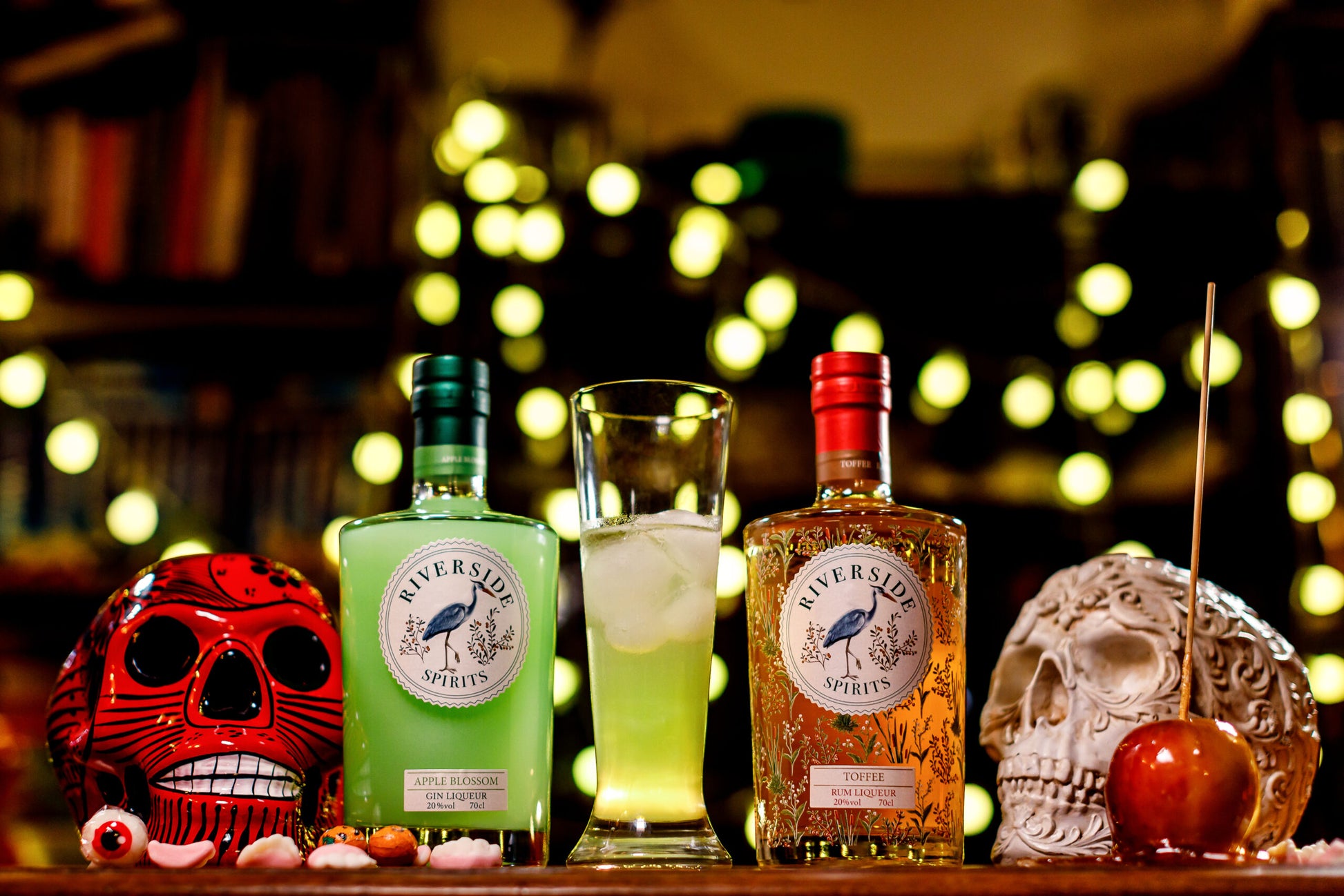 Two bottles of Riverside Spirits (Apple Blossom Gin Liqueur and Toffee Rum Liqueur) with a cocktail on a bar counter, surrounded by decorative skulls and fruits for Halloween. 