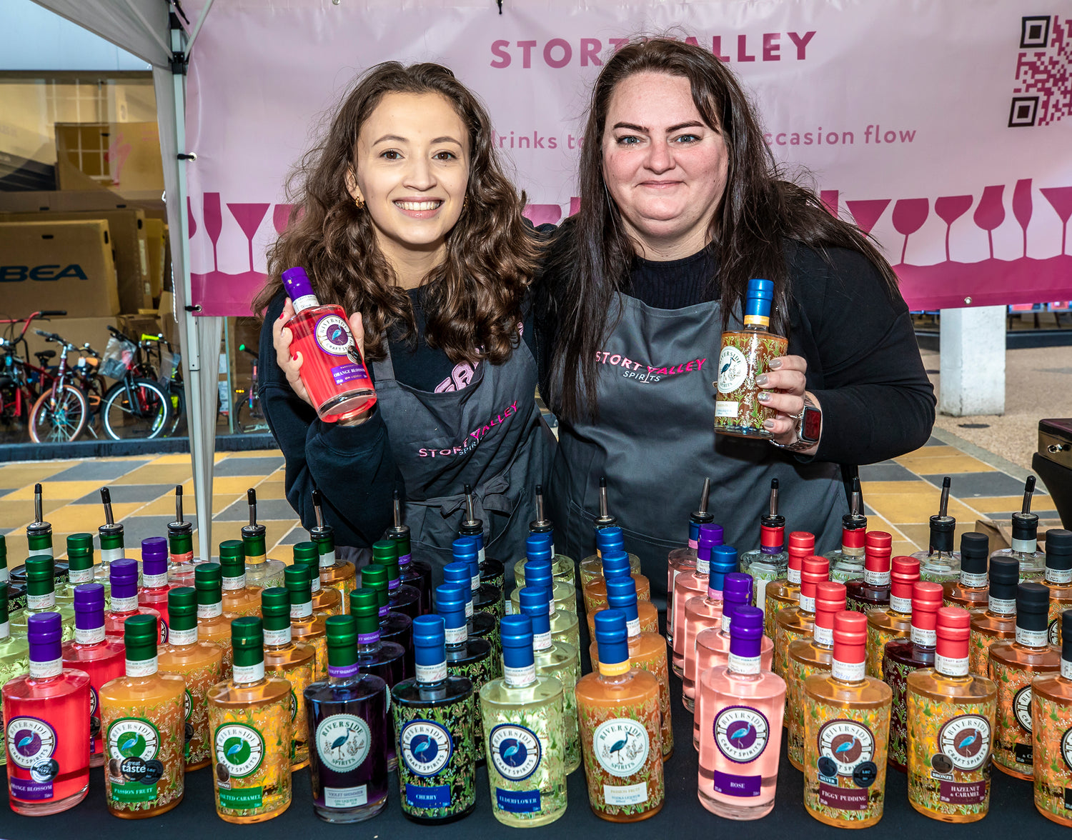 Two women standing behind a table displaying various Riverside spirits with colourful labels at an outdoor event.