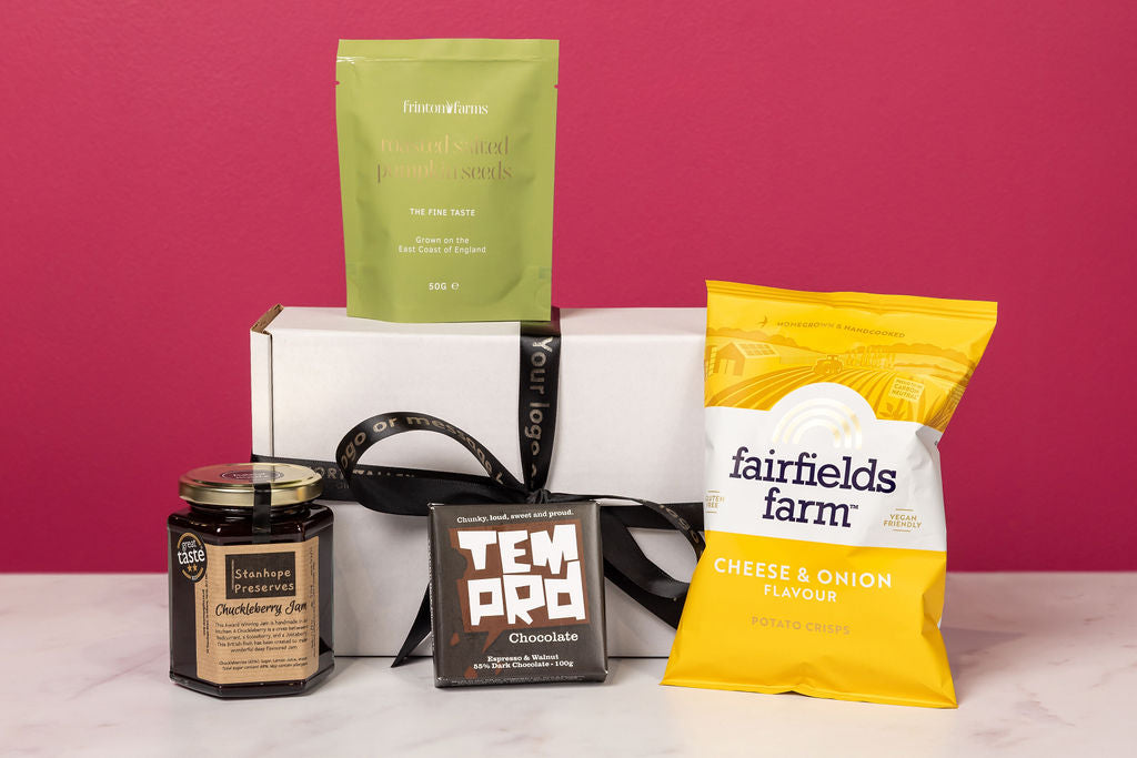 The Stort Valley Gifting celebration gift box including: Assorted products including a green tea package, a yellow cheese and onion snack bag, a black chocolate bar, and a white box on a pink background.