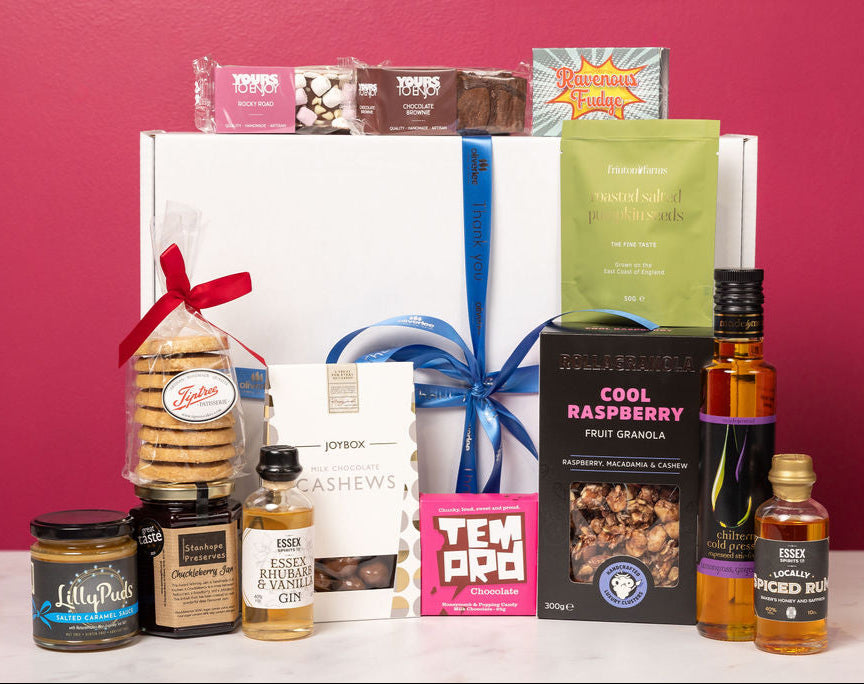 Gift basket with various items including snacks, drinks, and a box of chocolates against a pink background.