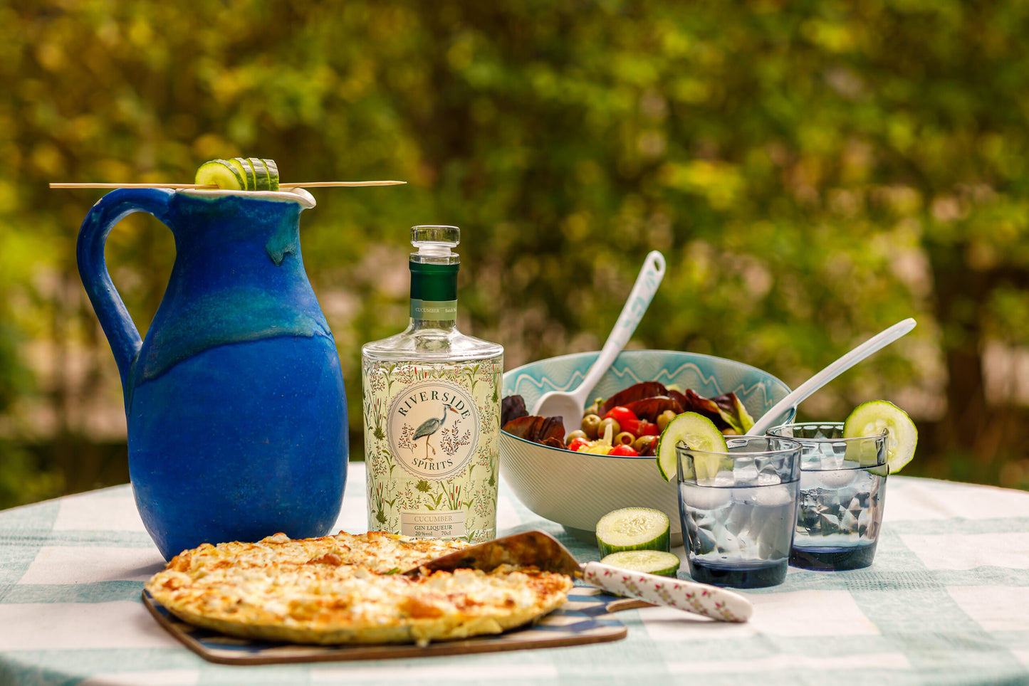 Outdoor setting with a table featuring a pizza, bottle of Riverside Spirits cucumber gin liqueur, and various items on a checkered table clothe