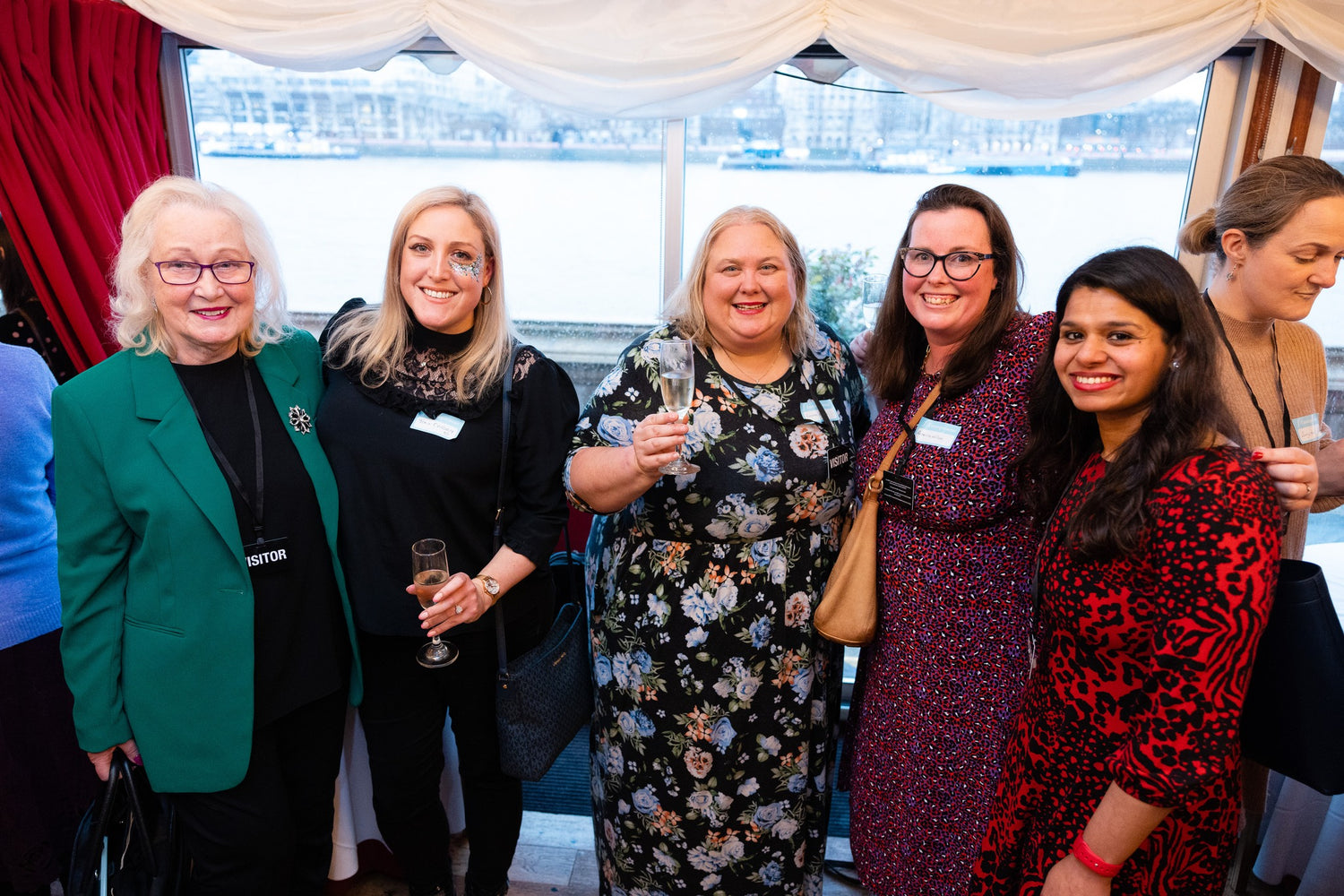 Group of women posing together at a business awards event with a window and red curtain in the background