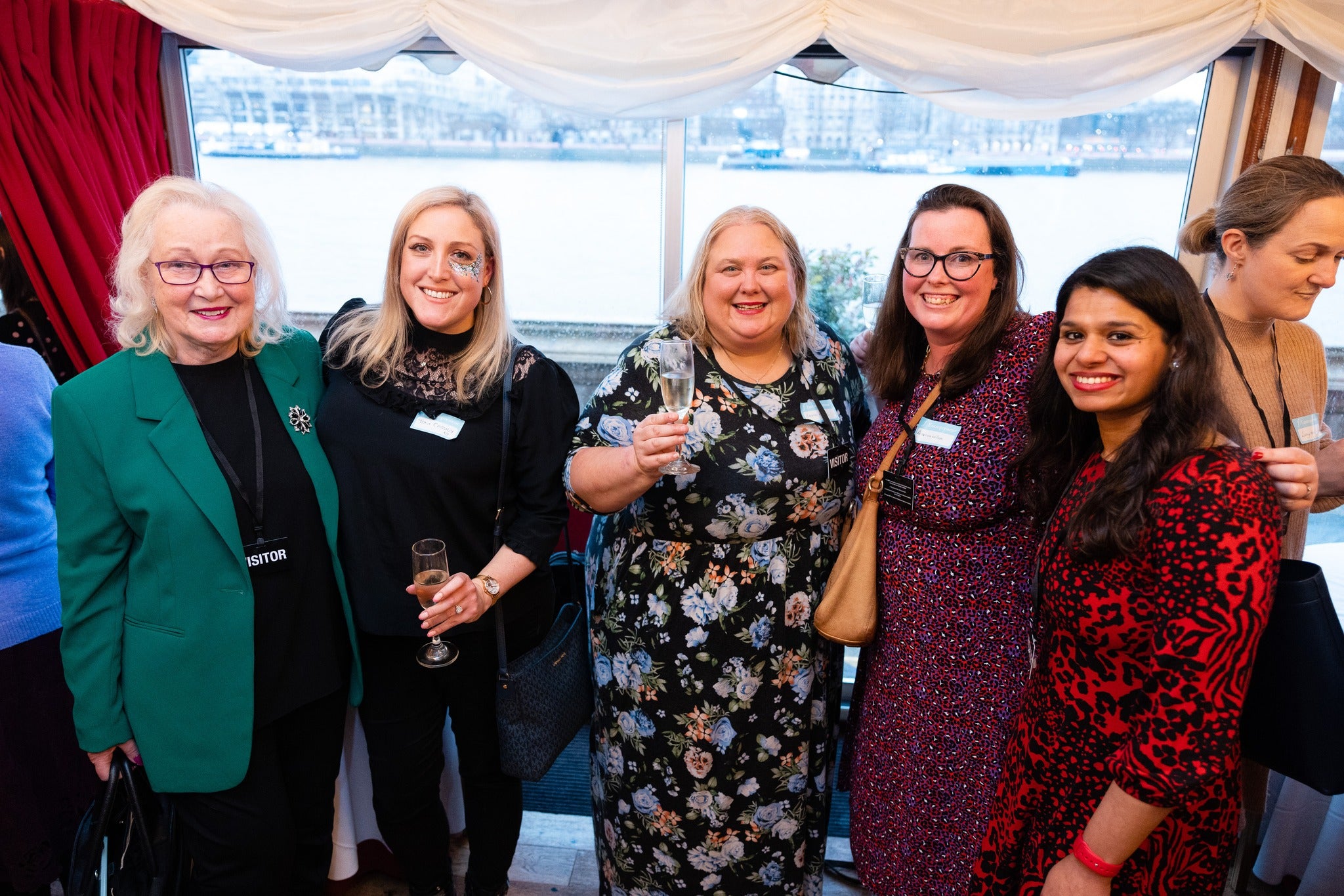 Group of women posing together at a business awards event with a window and red curtain in the background