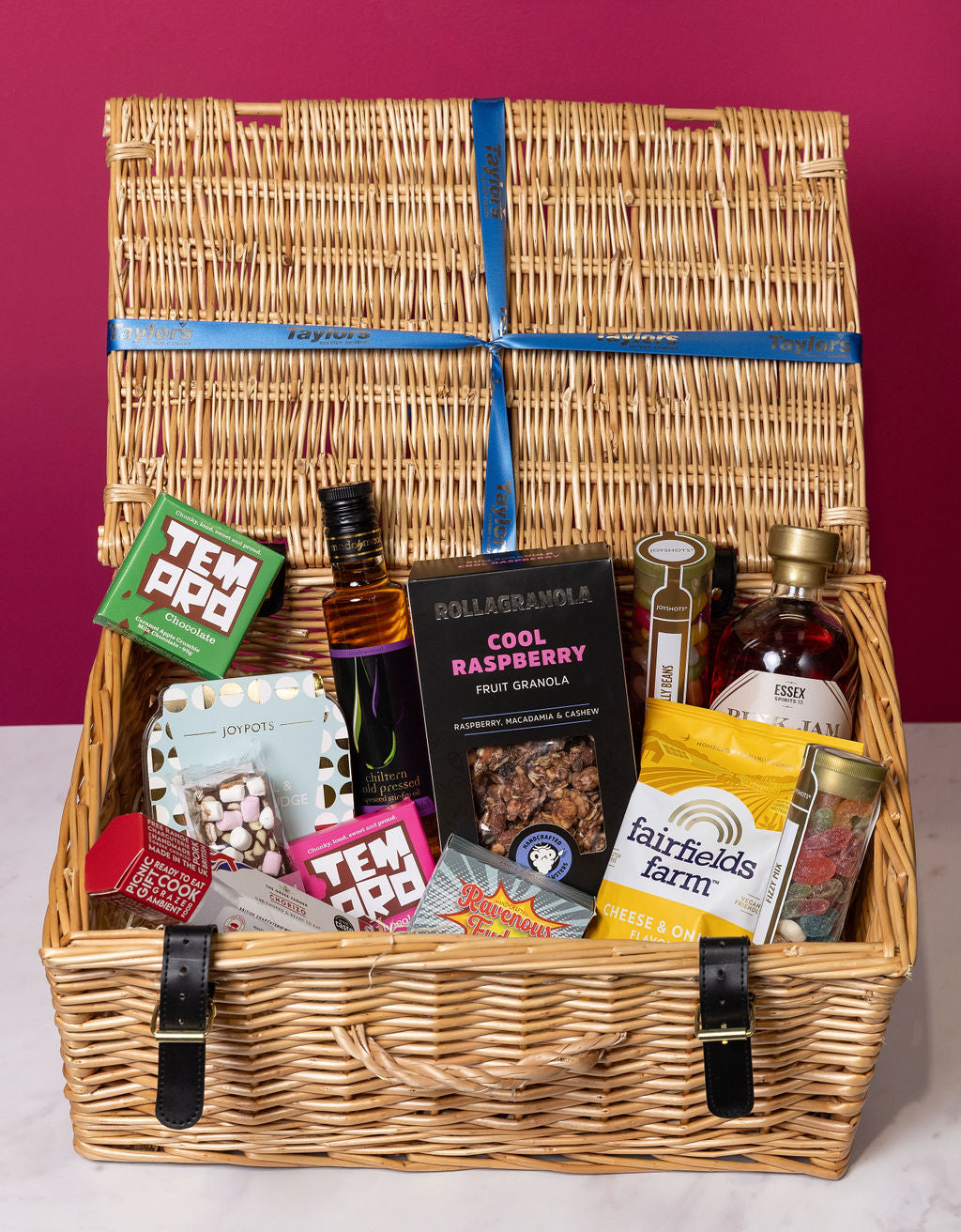 Wicker gift basket with various items including a bottle of alcohol and snacks on a red background