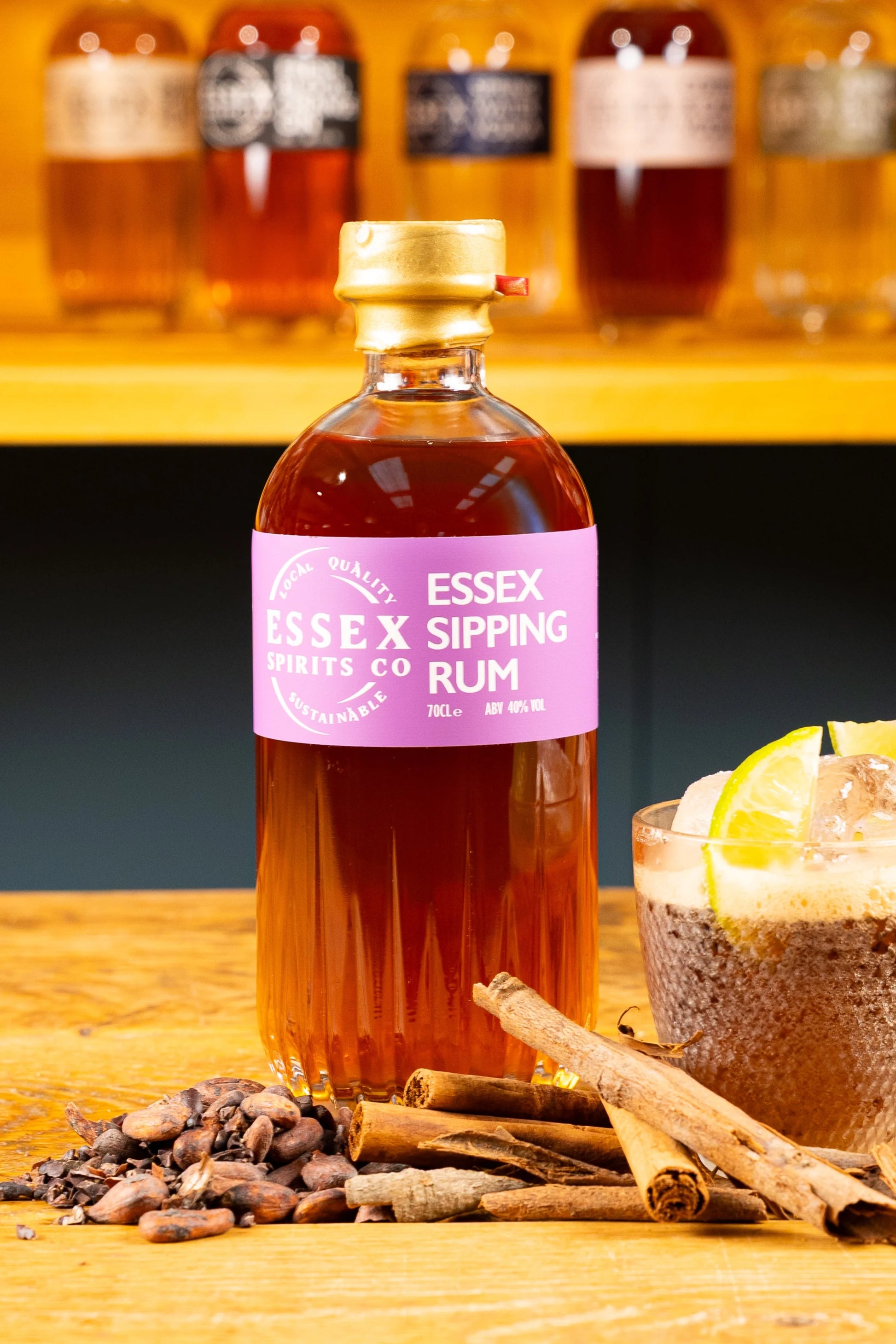 Bottle of Essex Sipping Rum on a wooden surface with spices and a cocktail in the background available at Stort Valley Gifting
