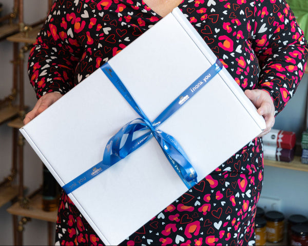 Person holding a personalised Stort Valley Gifting white gift box with a blue ribbon, wearing a colourful heart-patterned outfit.
